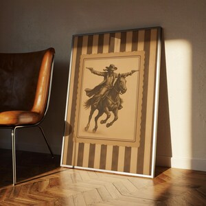 May include: Framed sepia-toned print of a cowboy on horseback, arms outstretched, holding two pistols. The artwork is set against a striped background with a silver frame. A brown leather chair sits to the left.