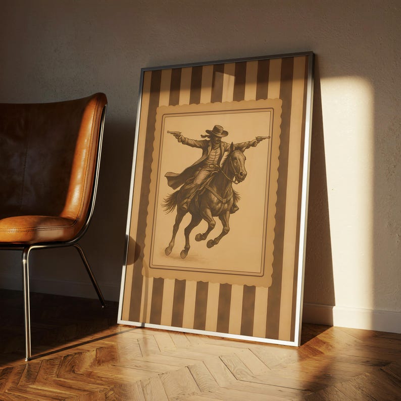 May include: Framed sepia-toned print of a cowboy on horseback, arms outstretched, holding two pistols. The artwork is set against a striped background with a silver frame. A brown leather chair sits to the left.