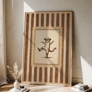 May include: A framed print featuring a sepia-toned illustration of a cat wearing a cowboy hat and boots, striking a dancing pose. The print is set against a background of vertical brown and beige stripes, with a light wooden frame.