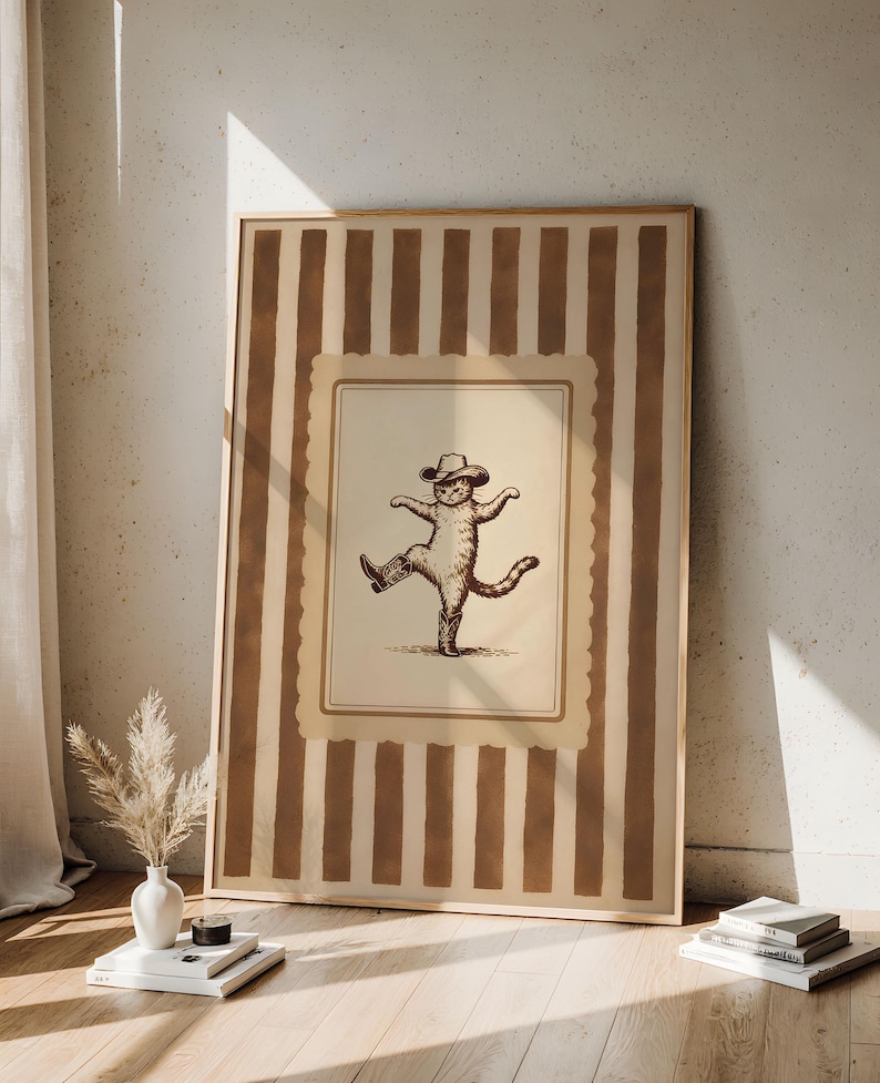 May include: A framed print featuring a sepia-toned illustration of a cat wearing a cowboy hat and boots, striking a dancing pose. The print is set against a background of vertical brown and beige stripes, with a light wooden frame.
