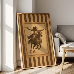 May include: Framed sepia-toned print of a cowboy on horseback, guns drawn. The artwork is set against a striped background with a decorative border, encased in a wooden frame. The print leans against a white wall.