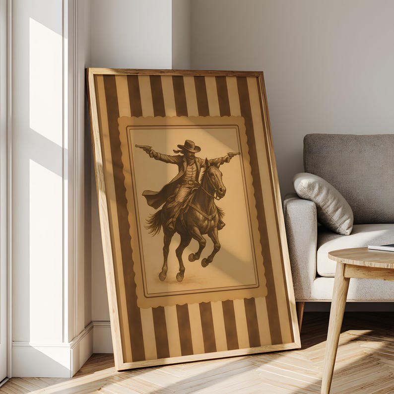 May include: Framed sepia-toned print of a cowboy on horseback, guns drawn. The artwork is set against a striped background with a decorative border, encased in a wooden frame. The print leans against a white wall.