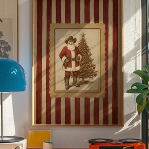 May include: Framed print featuring a cowboy Santa Claus with a Christmas tree. The artwork is set against a red and white striped background. The image is in a wooden frame and is displayed on a white wall.