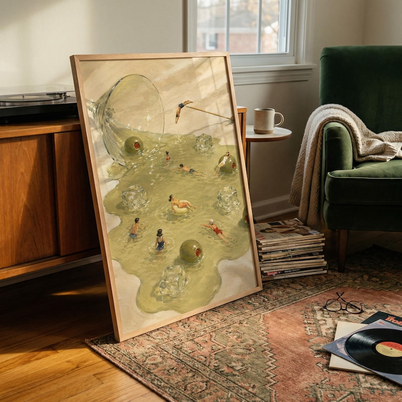 May include: A framed art print featuring a surreal illustration of a spilled martini glass. The liquid forms a pool containing small figures swimming, floating on ice cubes, and diving, alongside large green olives in a light olive-green liquid.