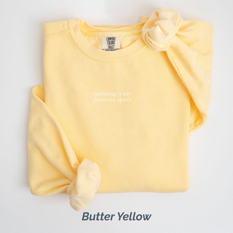 May include: Butter yellow sweatshirt with the text "mahjong is my favorite sport" in white. The sweatshirt has a crew neck and long sleeves. The brand label "Comfort Colors" is visible on the neckline.