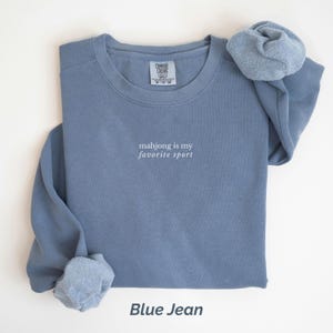 May include: A blue jean-colored sweatshirt with the text "mahjong is my favorite sport" in white. The sweatshirt has a crew neck and long sleeves. A matching scrunchie is placed on top of the sweatshirt.