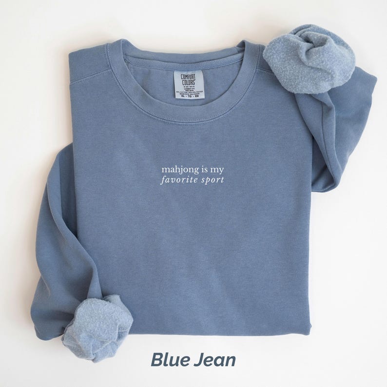 May include: A blue jean-colored sweatshirt with the text "mahjong is my favorite sport" in white. The sweatshirt has a crew neck and long sleeves. A matching scrunchie is placed on top of the sweatshirt.