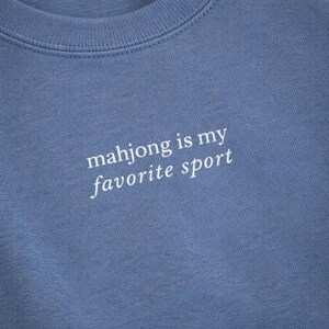 May include: A close-up of a blue crew neck sweatshirt. The white text on the front reads "mahjong is my favorite sport". The fabric has a soft texture. The sweatshirt is a solid color.
