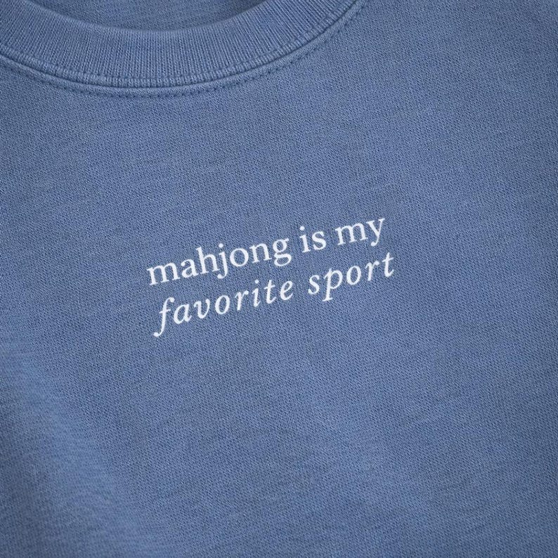 May include: A close-up of a blue crew neck sweatshirt. The white text on the front reads "mahjong is my favorite sport". The fabric has a soft texture. The sweatshirt is a solid color.