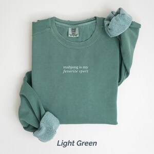 May include: Light green sweatshirt with the text "mahjong is my favorite sport" in white. The sweatshirt has a crew neck and long sleeves with a light blue fleece lining at the cuffs. The words "Light Green" are at the bottom.