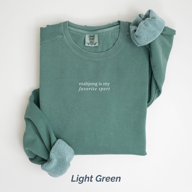 May include: Light green sweatshirt with the text "mahjong is my favorite sport" in white. The sweatshirt has a crew neck and long sleeves with a light blue fleece lining at the cuffs. The words "Light Green" are at the bottom.