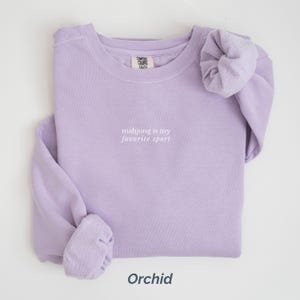 May include: Orchid-colored sweatshirt with the text "mahjong is my favorite sport" in white. The sweatshirt is folded neatly, with the sleeves and cuffs visible. The word "Orchid" is printed at the bottom.