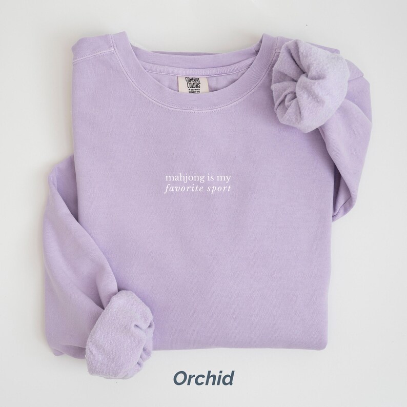 May include: Orchid-colored sweatshirt with the text "mahjong is my favorite sport" in white. The sweatshirt is folded neatly, with the sleeves and cuffs visible. The word "Orchid" is printed at the bottom.