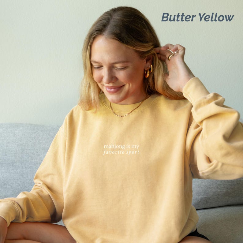 May include: Butter yellow sweatshirt with the text "mahjong is my favorite sport" in white. The sweatshirt has long sleeves and a crew neck. The model is wearing gold hoop earrings and a gold necklace.
