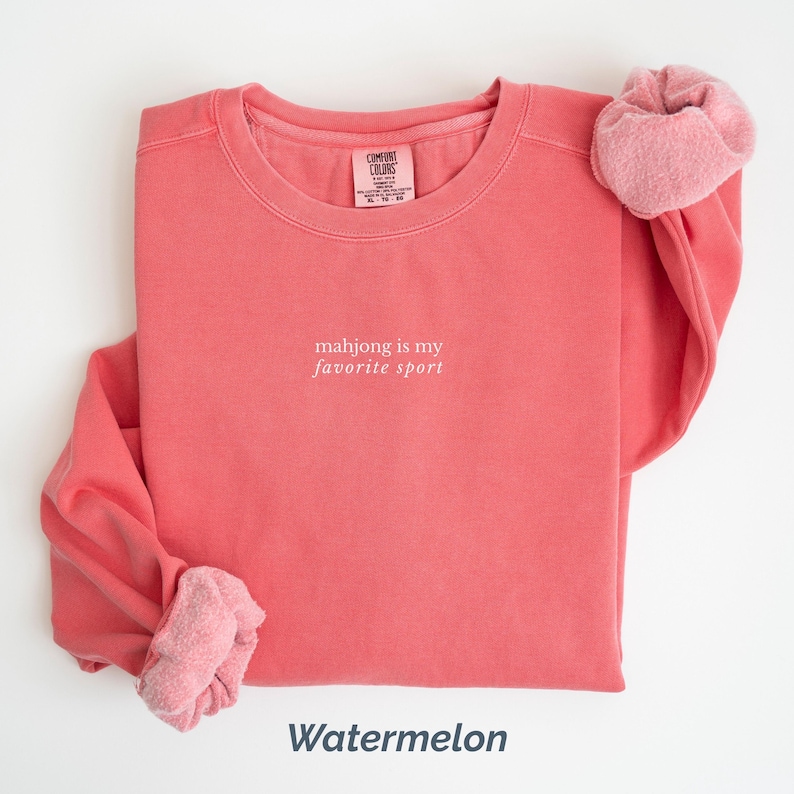 May include: A watermelon-colored sweatshirt with the text "mahjong is my favorite sport" in white. The sweatshirt has a crew neck and long sleeves, with a pink scrunchie on the sleeve. The word "Watermelon" is at the bottom.