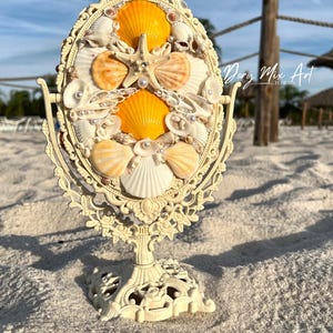 May include: An ornate, cream-colored vanity mirror decorated with seashells and a starfish. The mirror's oval frame is embellished with various seashells, pearls, and a central starfish. The mirror is on a decorative stand, and the text "Dory Mix Art - FLORIDA" is visible.