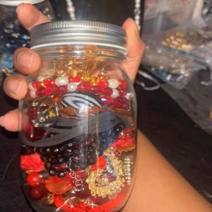 Puede incluir: Un tarro de cristal transparente lleno de varias joyas. El tarro contiene cuentas, pulseras y otros accesorios rojos, negros y dorados. El tarro tiene una tapa plateada y lo sostiene una persona.