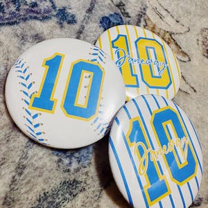 Puede incluir: Tres botones redondos con el número 10 en azul y amarillo. Uno está diseñado como una pelota de béisbol, los otros dos tienen un fondo a rayas. La palabra "Janeway" está escrita en dos de los botones.