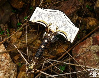 May include: A decorative hammer with a white head featuring an intricate design and gold accents. The handle is wrapped in brown leather and gold-colored metal. The hammer is set against a natural background of rocks and twigs.