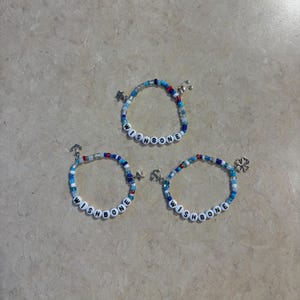 Puede incluir: Tres pulseras de cuentas con la palabra "WISHBONE" escrita con cuentas de letras blancas. Las pulseras presentan cuentas azules, rojas y transparentes, con dijes que incluyen estrellas, anclas y un trébol de cuatro hojas. Las pulseras se exhiben sobre una superficie neutra.