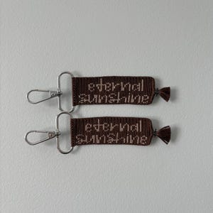 May include: Two rectangular brown woven keychains featuring the text "eternal sunshine" embroidered in a light beige thread. Each keychain is attached to a silver-toned metal lobster clasp and features a small brown tassel at the opposite end.