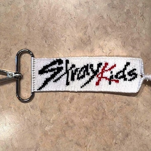 May include: White woven keychain with the text "Stray Kids" in black and red. The keychain has a silver-colored clasp and a white tassel. The text is embroidered on the white fabric.