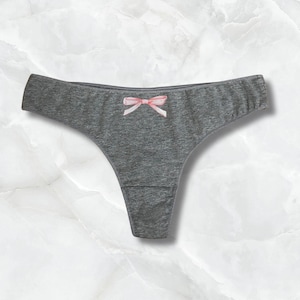 Puede incluir: Tanga gris con un pequeño lazo rosa en la parte delantera. La ropa interior está hecha de una tela suave y texturizada. El fondo es un patrón de mármol.