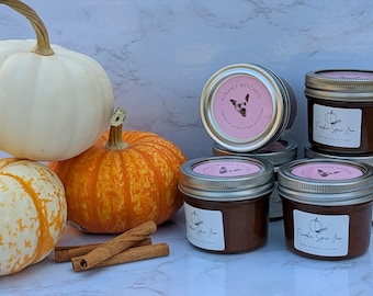 May include: A display of pumpkins, cinnamon sticks, and jars of pumpkin spice jam. The pumpkins are white, orange, and yellow. The jam jars have pink labels with a pumpkin illustration and the text "Pumpkin Spice Jam".