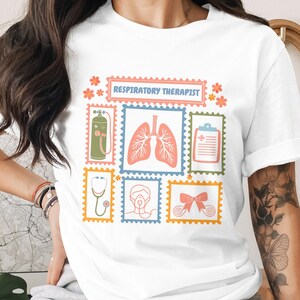May include: White t-shirt featuring a colorful design with the text "RESPIRATORY THERAPIST". The design includes illustrations of lungs, a stethoscope, an oxygen tank, and other medical-related images. The shirt is made of a soft material.