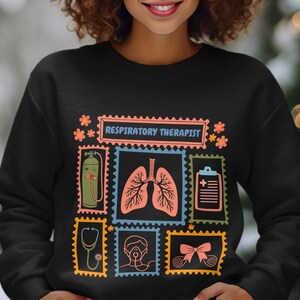 May include: Black sweatshirt with a colorful design featuring the text "RESPIRATORY THERAPIST" and illustrations of medical equipment and lungs. The design includes a stethoscope, oxygen tank, and a medical chart.