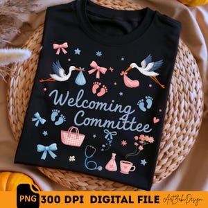 Op de afbeelding: Zwart T-shirt met een baby shower ontwerp. Het shirt heeft ooievaars die baby's dragen, strikken, babyvoetjes en de woorden "Welcoming Committee". Extra afbeeldingen zijn een stethoscoop, een handtas en een kopje koffie.