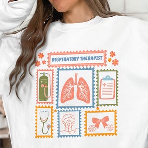 May include: White sweatshirt featuring a colorful graphic design with the text "Respiratory Therapist." The design includes illustrations of lungs, a stethoscope, and an oxygen tank, among other medical-related images.