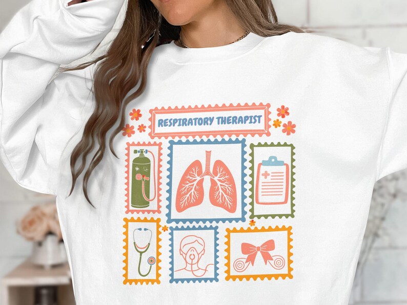 May include: White sweatshirt featuring a colorful graphic design with the text "Respiratory Therapist." The design includes illustrations of lungs, a stethoscope, and an oxygen tank, among other medical-related images.