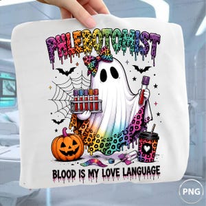 Puede incluir: Una camiseta blanca con un fantasma de dibujos animados que lleva un lazo con estampado de leopardo de colores, sosteniendo un gradilla de tubos de ensayo y un tubo. El diseño incluye una calabaza, telaraña, murciélagos y un vaso con el texto PHLEBOTOMIST y BLOOD IS MY LOVE LANGUAGE.