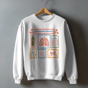 May include: White sweatshirt featuring a colorful design with the text "RESPIRATORY THERAPIST". The design includes illustrations of medical equipment and the lungs, set within postage stamp-like frames. The sweatshirt is hanging on a wooden hanger.