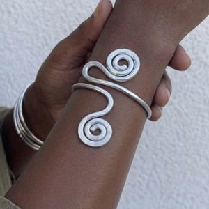 Op de afbeelding: Een zilverkleurige metalen manchetarmband met een doorlopend draadontwerp dat twee opvallende, symmetrische spiralen vormt aan de buitenzijde. De armband heeft een gladde, gepolijste afwerking en wikkelt zich om de pols met een open structuur.