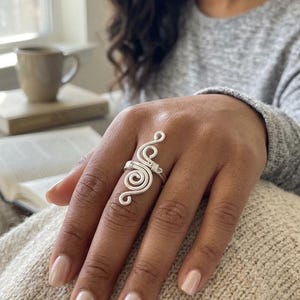 Könnte beinhalten: Ein silberfarbener, drahtgewickelter Ring mit einem markanten Spiraldesign, das sich vertikal entlang des Fingers erstreckt. Der Metalldraht ist in der Mitte zu einem kreisförmigen Muster gewickelt, mit kleineren dekorativen Schlaufen an den Enden.