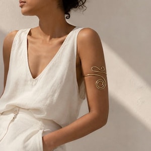 Pode incluir: Um bracelete de metal em tom dourado com um design de espiral dupla. A peça de joalharia é usada na parte superior do braço e consiste num fio contínuo que forma duas bobinas circulares distintas ligadas por uma barra horizontal.