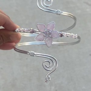 Puede incluir: Un brazalete de alambre plateado con una flor central de cinco pétalos hecha de piedra rosa claro. El alambre tiene dos extremos en espiral y está decorado con pequeñas cuentas rosas translúcidas a lo largo de la banda.