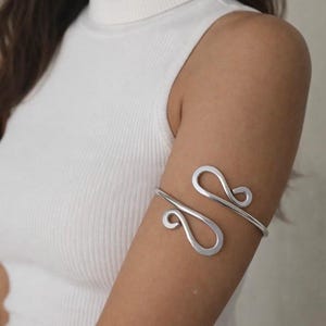Peut inclure: Un bracelet de bras argenté avec un design ouvert et fluide. Le bracelet présente deux éléments décoratifs incurvés et s'enroule autour du haut du bras. Le bijou est porté sur un haut sans manches blanc.
