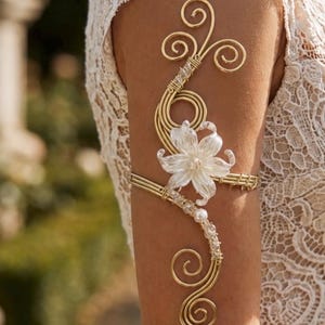 Op de afbeelding: Een bovenarmband van goudkleurig draad met een centraal wit bloemaccent, kleine doorzichtige kralen en een enkele witte parel. De metaaldraad is gevormd in decoratieve krullen die verticaal langs de bovenarm lopen voor een gestructureerd ontwerp.