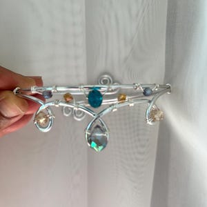 Può includere: Una corona in filo metallico argentato con una perla ovale color ottanio al centro, un cristallo iridescente a goccia pendente e perline decorative più piccole nei toni dell'ambra e del grigio. Il filo forma spirali e riccioli decorativi.