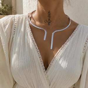 Może przedstawiać: Srebrzysty metalowy naszyjnik typu choker o otwartej konstrukcji z przodu, z dwoma płaskimi końcami skierowanymi w dół. Naszyjnik spoczywa na szyi, prezentując gładkie, polerowane wykończenie, które odbija światło na białej, fakturowanej tkaninie.