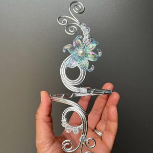 Pode incluir: Um acessório de cabelo floral em metal prateado. O design apresenta uma flor azul translúcida com um centro de pérola, cristais transparentes e detalhes de arame em espiral. O acessório é segurado em uma mão contra um fundo escuro.