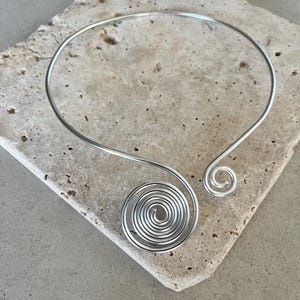 Op de afbeelding: Een zilverkleurige metalen choker met een cirkelvormig draadontwerp. De ketting heeft aan één uiteinde een grote, meerlaagse spiraalvormige krul en aan het andere uiteinde een kleinere, enkele spiraal, verbonden door een dunne, flexibele metalen band.