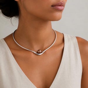 Peut inclure: Un collier ras-du-cou en métal argenté composé d'un fil incurvé et lisse qui s'affine vers le centre. Une perle sphérique unique, polie et de couleur brun foncé, est maintenue fermement dans le berceau central de la structure métallique.