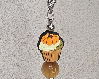Könnte beinhalten: Silberfarbener Kettenanhänger mit einem Cupcake-Anhänger mit einem Kürbis darauf. Der Cupcake hat orange, weiße und braune Details. Unter dem Anhänger befindet sich eine runde, bernsteinfarbene Perle.