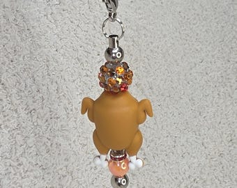 Op de afbeelding: Zilverkleurige ketting met een bruine kalkoen hanger, witte voetjes en een decoratief oranje en goudkleurig kralenaccent. De hanger hangt aan een zilveren ketting met een karabijnsluiting.
