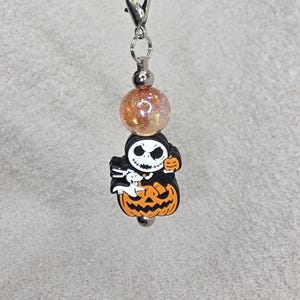 May include: A Halloween charm featuring a Jack Skellington figure on an orange pumpkin. The charm has a silver clasp and a shimmering orange bead. The Jack Skellington figure is black and white with a small ghost.