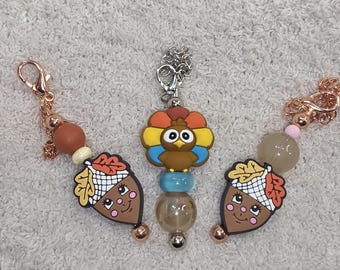 Op de afbeelding: Drie Thanksgiving-thema sleutelhangers. Eén heeft een kalkoenontwerp met een kleurrijke waaier, en de andere twee hebben eikelbedels met lachende gezichten. Elke sleutelhanger heeft een andere kraal- en kettingkleur.